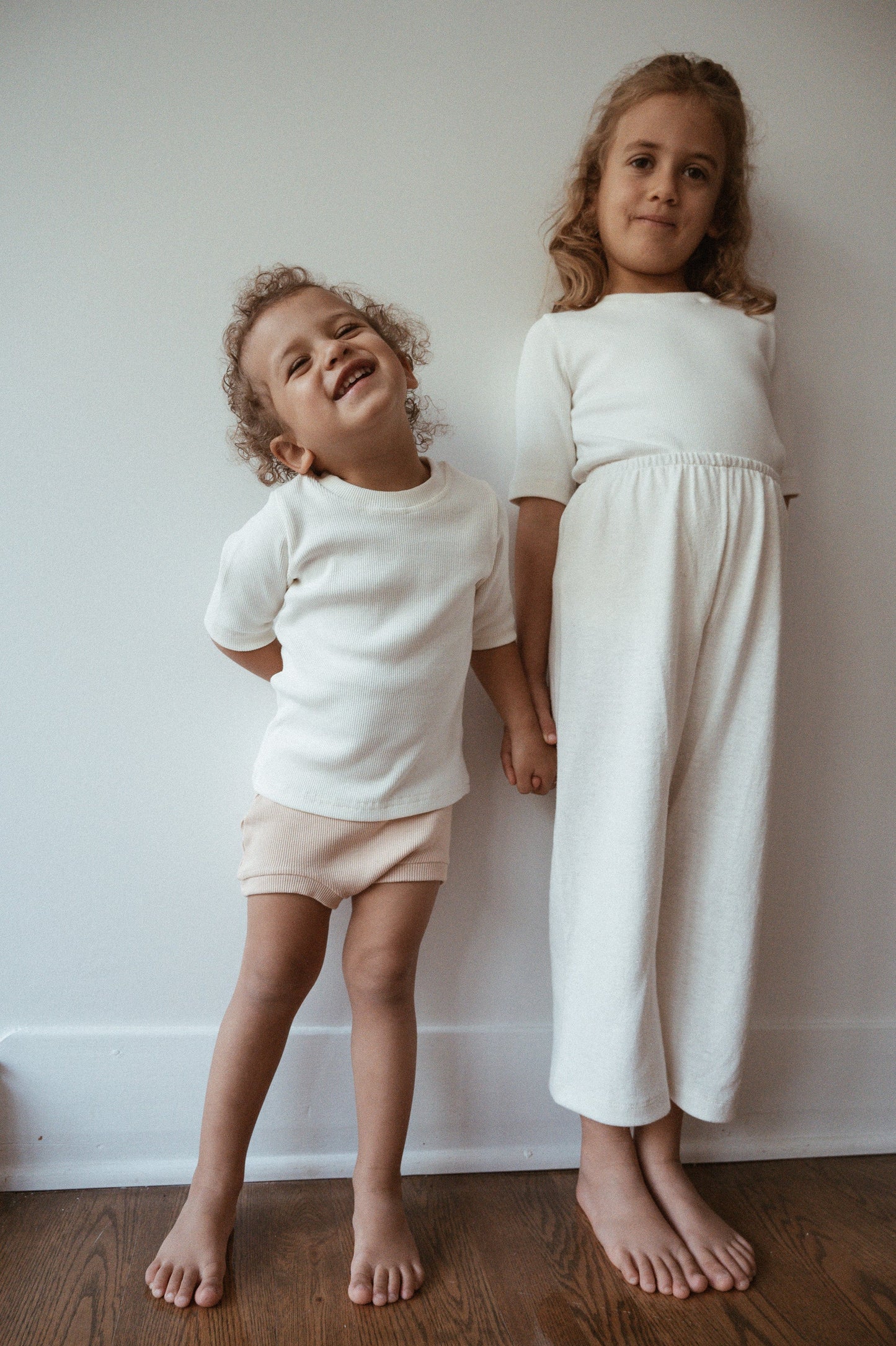 Two young children stand barefoot against a plain white wall. The younger child on the left wears a white t-shirt and light shorts, smiling and tilting their head. The older child on the right wears a white long-sleeve top and Resort Pant in Hemp from cabane childrenswear, standing straight and looking at the camera.