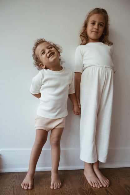 Two young children stand barefoot against a plain white wall. The younger child on the left wears a white t-shirt and light shorts, smiling and tilting their head. The older child on the right wears a white long-sleeve top and Resort Pant in Hemp from cabane childrenswear, standing straight and looking at the camera.