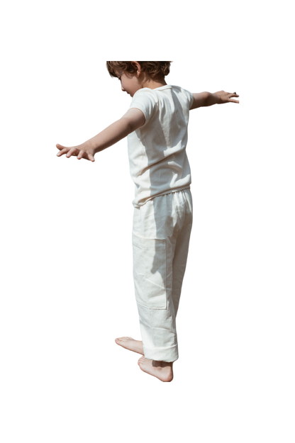 A child in a white outfit stands with arms outstretched, appearing to balance or play, wearing the handmade Pantalon D'Artiste in Hemp Twill by cabane childrenswear. The plain background highlights the child's playful pose and emphasizes the charm of this ethical children's wear.