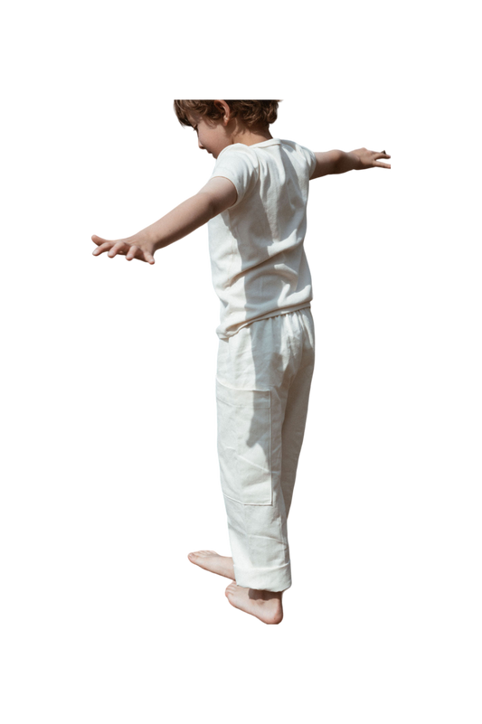 A young child, dressed in an ethically handmade light-colored PANTALON D'ARTISTE hemp twill outfit by cabane childrenswear, stands on their tiptoes with arms outstretched, facing away from the camera. The background is removed, giving the image a cut-out look.