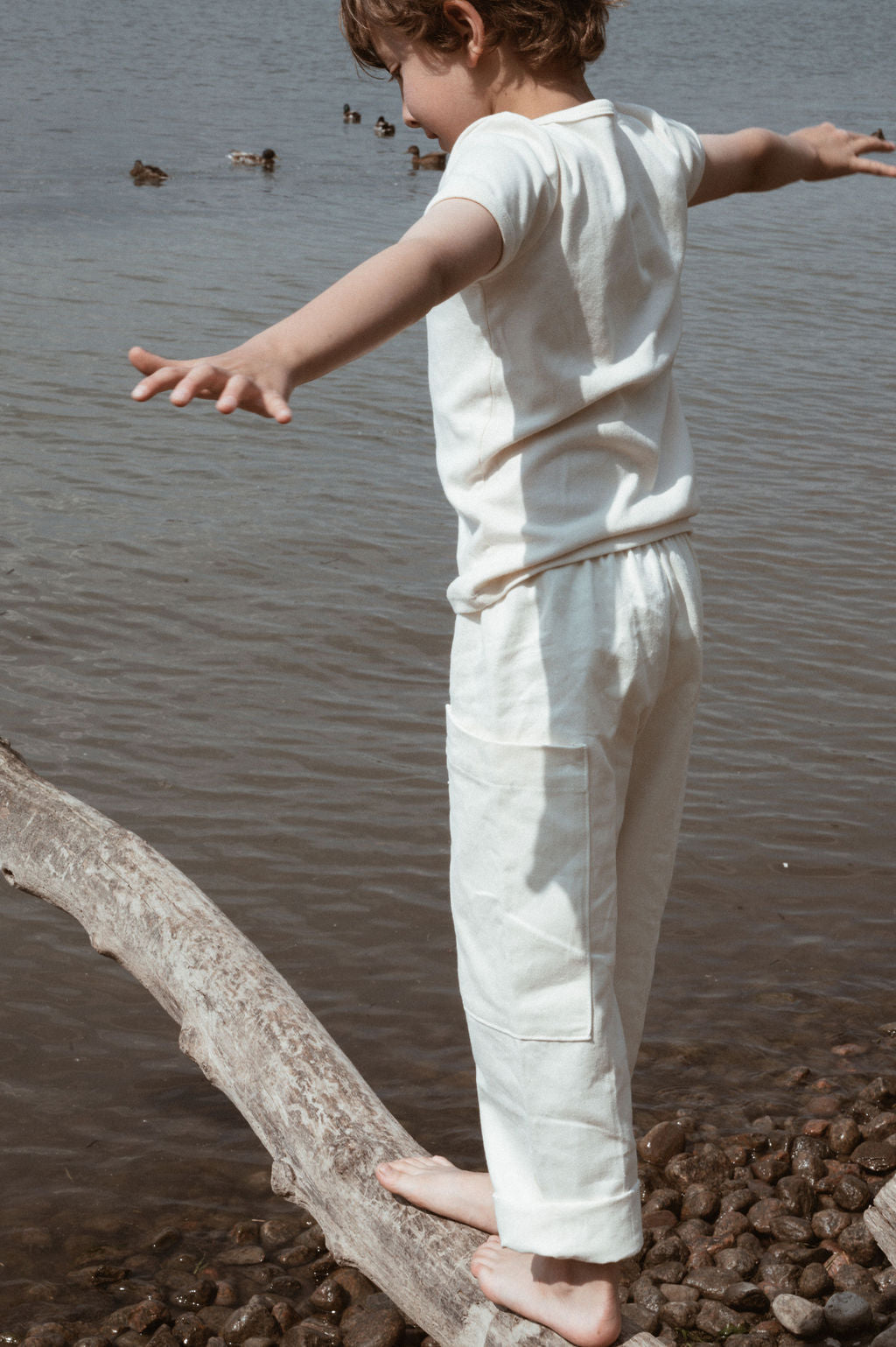 A child in cabane childrenswear's Pantalon D'Artiste in Hemp Twill balances on a log over a pebbly lake shore. Wearing a white t-shirt, they extend their arms while watching ducks swim in the water.