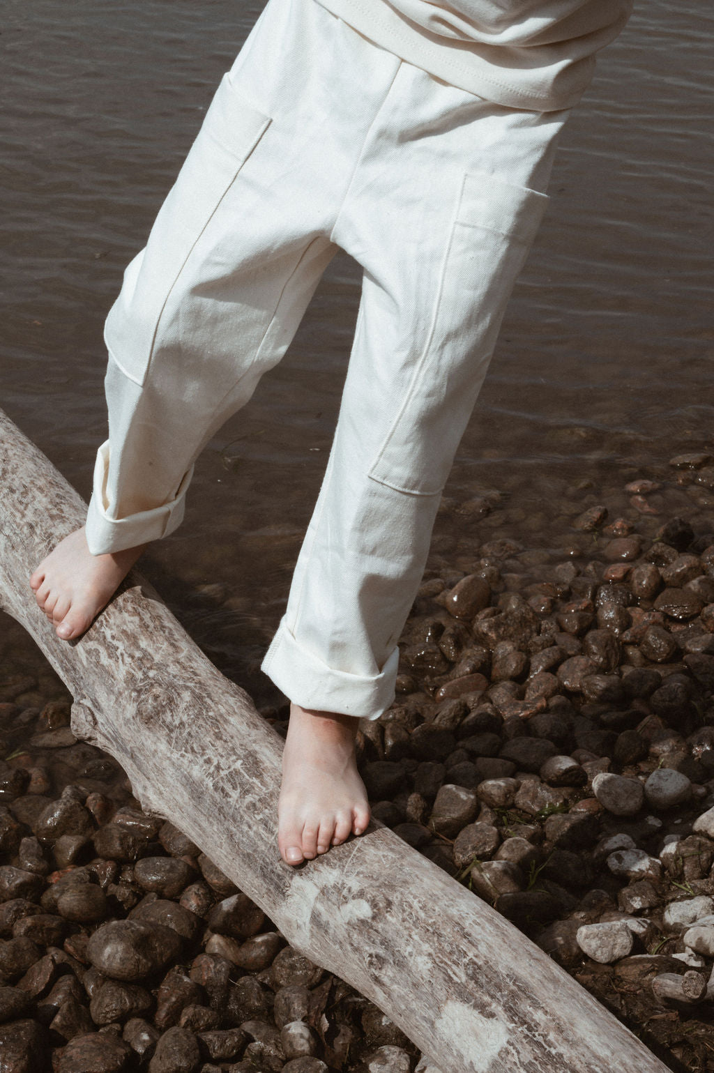 A person walks barefoot on a tree trunk above a rocky surface by the water, their rolled-up white pants resembling cabane childrenswear's Pantalon D'Artiste in Hemp Twill, known for its ethical design and flexible fit.