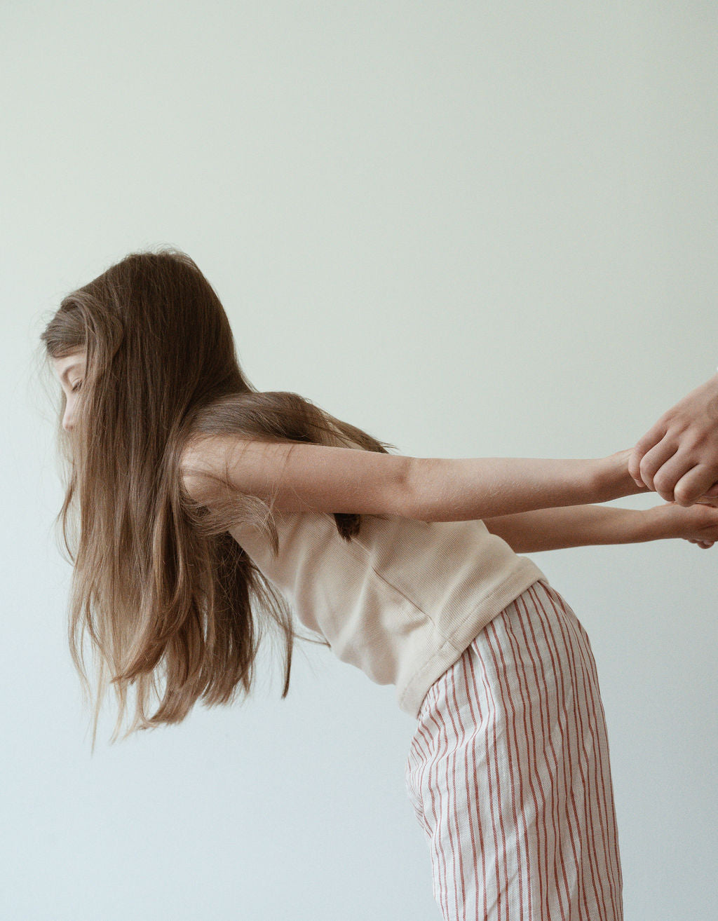 A young girl with long hair, dressed in a sleeveless top and a pair of PER SE PANT loom linen from cabane childrenswear, is leaning forward while holding hands with another person who is out of the frame against a neutral background.