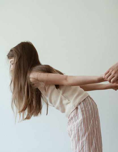 A young girl with long hair, dressed in a sleeveless top and a pair of PER SE PANT loom linen from cabane childrenswear, is leaning forward while holding hands with another person who is out of the frame against a neutral background.