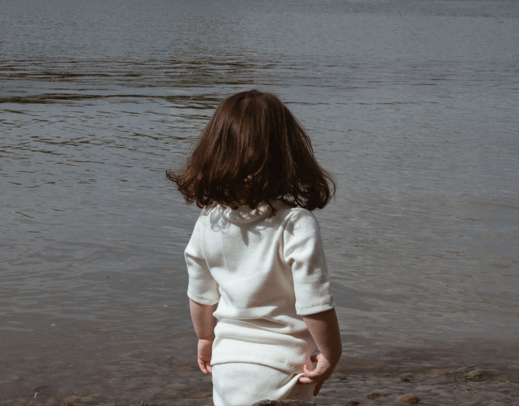 A young child with shoulder-length brown hair stands at the edge of a calm body of water, facing away from the camera. The child is wearing an ethically handmade CLASSIC SET baby + kid by cabane childrenswear with rib texture and gazes towards the water, where a few small stones and pebbles are visible on the ground.