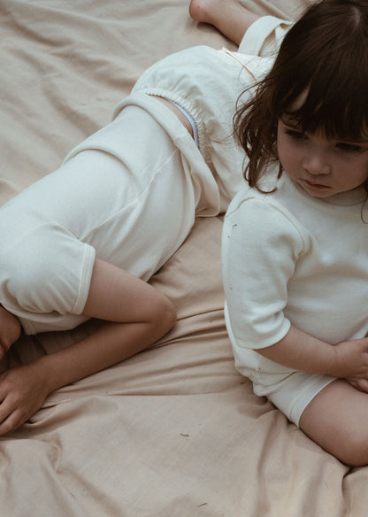 Two young children are lying on a beige fabric, both dressed in cream-colored outfits from the CLASSIC SET baby + kid by cabane childrenswear. One child lies on their stomach, facing away, while the other sits upright with a thoughtful expression. Their rib-textured outfits add to the scene's calm and intimate feel.