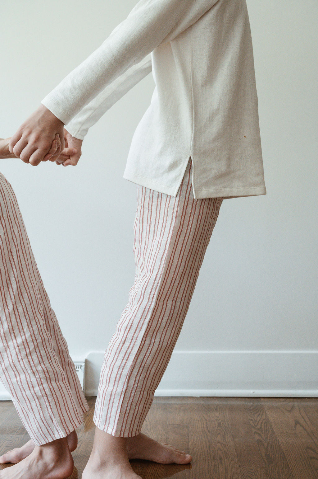 Two individuals are seen holding hands. One person is wearing a long-sleeved, off-white shirt paired with striped pajama pants cropped above the ankles. The other person, partially visible, is dressed in relaxed PER SE PANT loom linen by cabane childrenswear. Both outfits appear to be ethically made as they stand on a wooden floor.