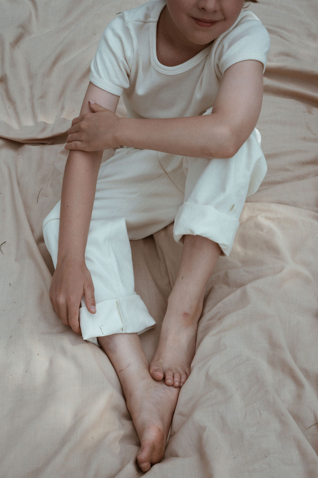 A child sits on a beige fabric, dressed in cabane childrenswear's sustainable Pantalon D'Artiste in Hemp Twill, featuring a white t-shirt and rolled-up white pants. They sit barefoot with one leg crossed over the other, hand resting on their knee, embodying a relaxed posture.