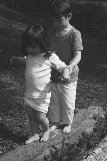 A black-and-white photo captures two young children walking barefoot on a log. The older child stands behind the younger one, holding their shoulders to help them balance as they carefully take steps. Both have serious expressions, concentrating on the activity, while dressed in attire from the cabane childrenswear CLASSIC SET baby + kid collection.