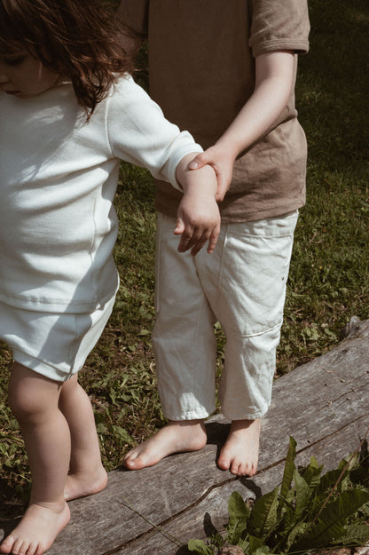 Two children walk barefoot on a fallen log in a grassy area. The child in the foreground, dressed in the cabane childrenswear CLASSIC SET baby + kid with its distinctive rib texture, holds hands with the child behind who wears a brown top and light-colored pants. They help each other balance under the sunny sky.