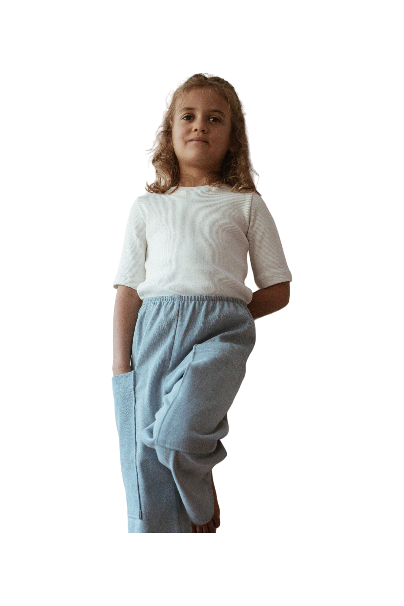 A young girl wearing a white shirt and the Pantalon D'Artiste in Denim from cabane childrenswear stands with one hand in her pocket, looking confidently at the camera. Her curly hair cascades gently around her shoulders, embodying the spirit of ethically handmade fashion in Calgary, Canada.