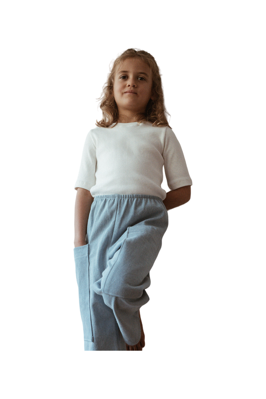 A young girl with curly hair stands confidently, wearing a white shirt and high-waist PANTALON D'ARTISTE denim pants from cabane childrenswear, with her hands in her pockets. The background is plain and dark, emphasizing her relaxed pose and casual attire.