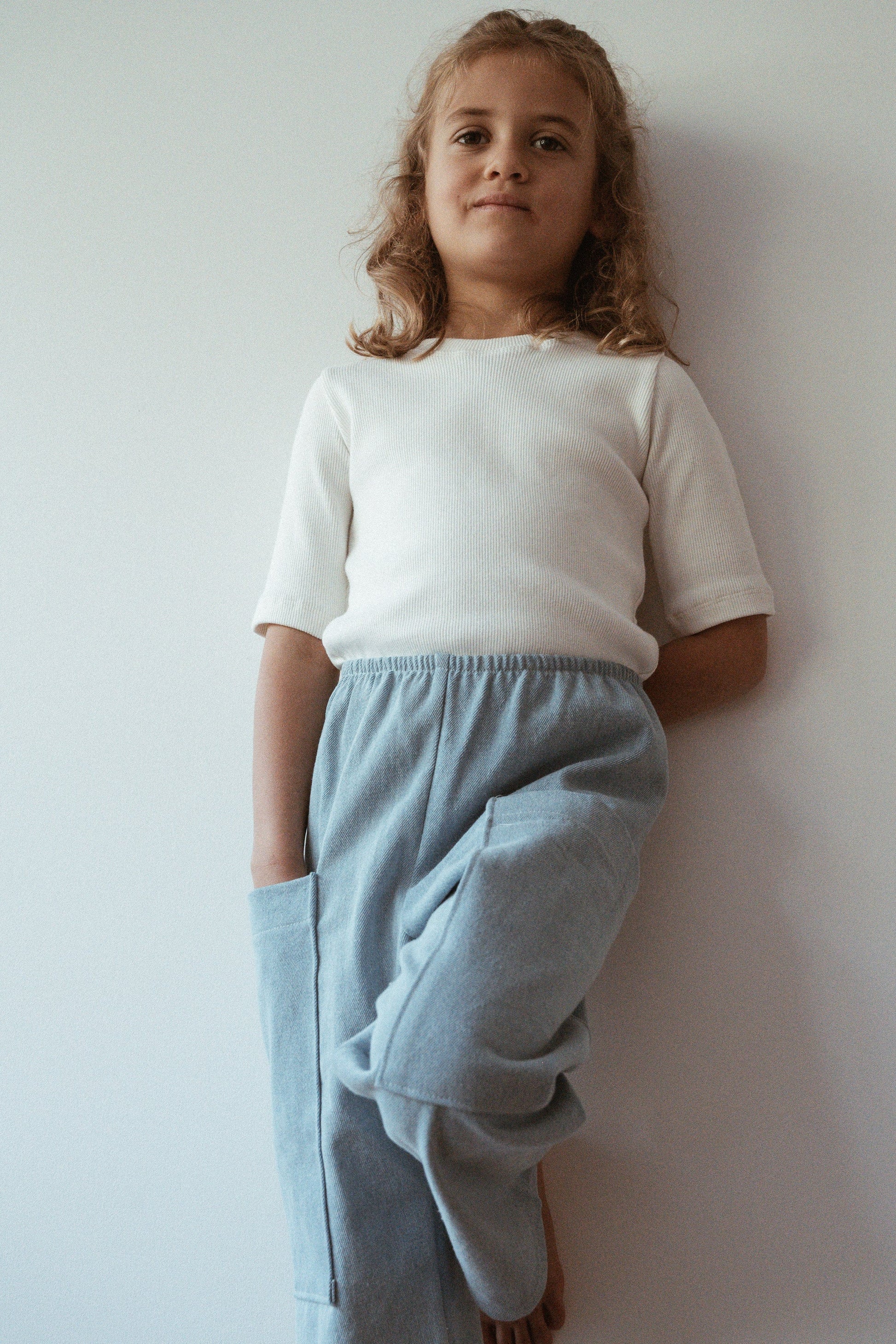 A young child with curly hair stands against a white wall, wearing a white shirt and the ethically handmade Pantalon D'Artiste in Denim from cabane childrenswear, based in Calgary, Canada. Their hands are tucked into their pockets, and they have a calm expression.