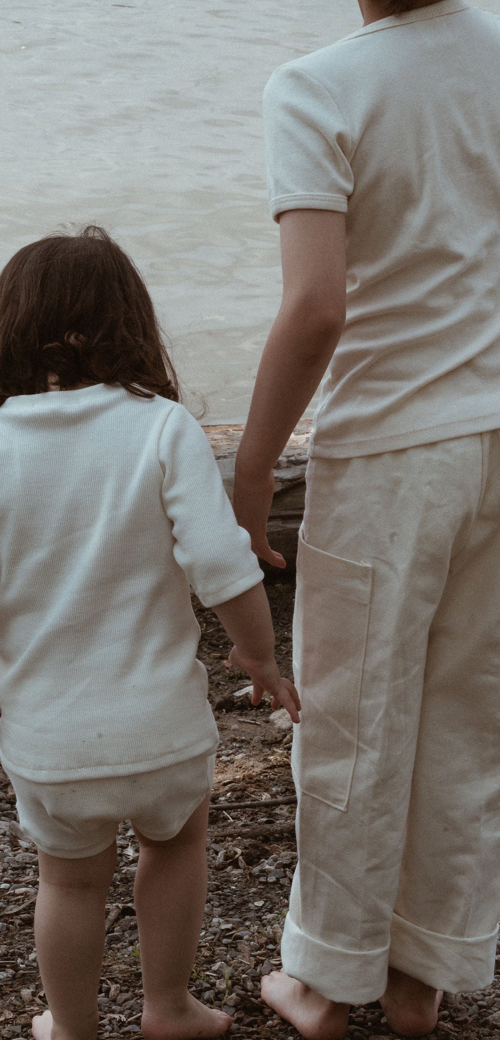 Two children in white clothing stand barefoot on a rocky shoreline, facing a body of water. The taller child wears cabane childrenswear's Pantalon D'Artiste in Hemp Twill, while the shorter one wears shorts. Their hands are close, as if about to hold hands—a perfect scene of ethical children's wear harmonizing with nature.