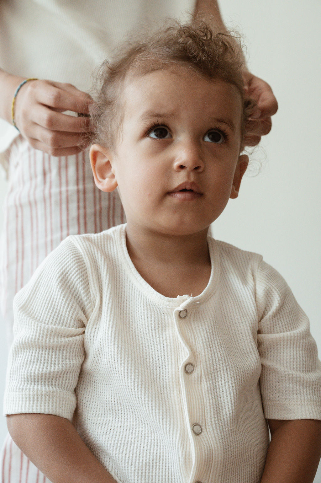 A young child with curly hair, dressed in a pre-order HIGH-NOON SUNSUIT from cabane childrenswear, looks upwards with a thoughtful expression. An adult behind them, only their hands visible, is gently fixing the child's hair. The background is softly lit and neutral.