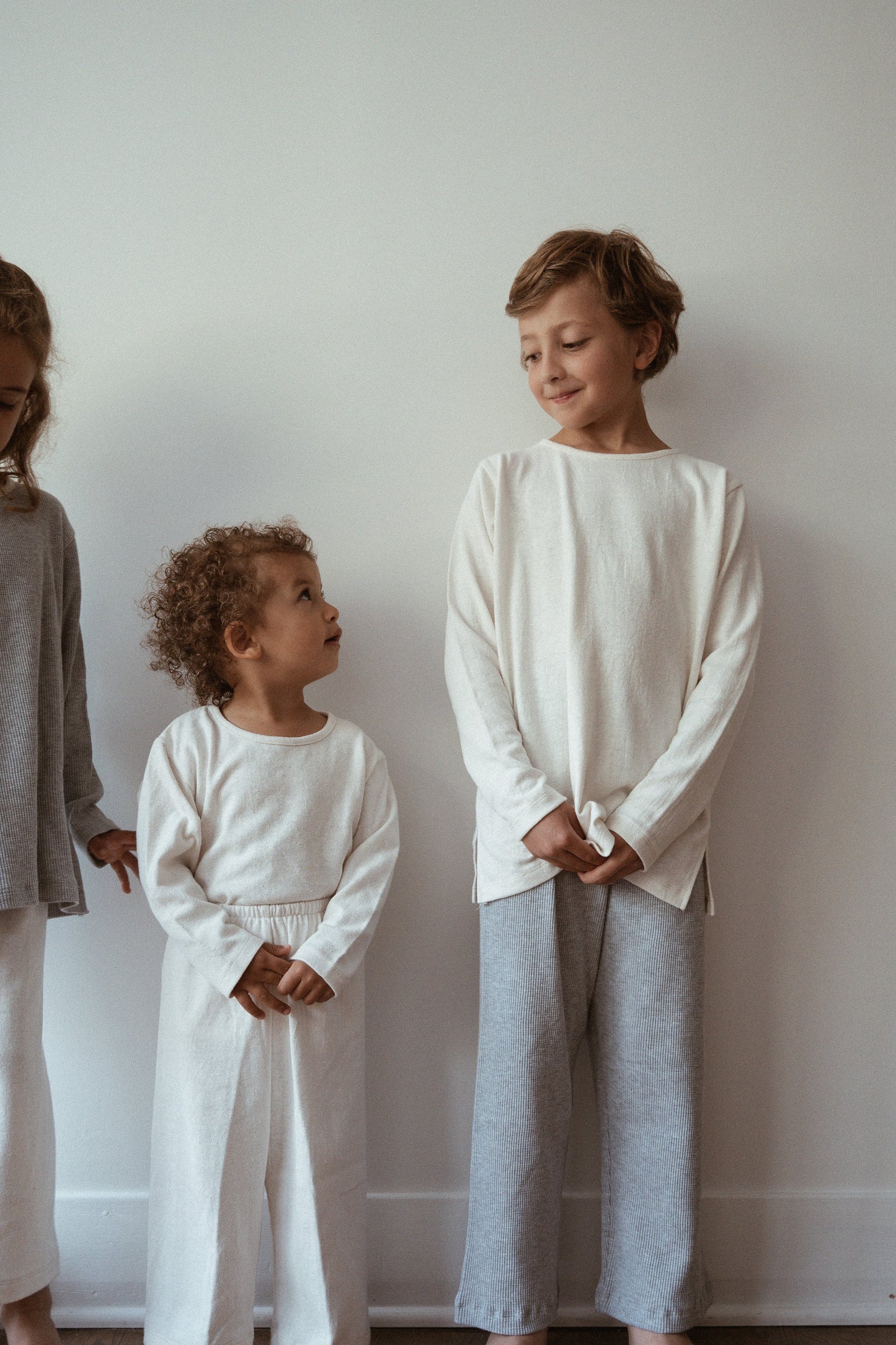 Three children stand in a row against a light-colored wall. The child on the left wears a gray top and white pants; the middle child, with curly hair, is dressed in all white and wears an adorable RESORT TOP hemp jersey handmade in Calgary by cabane childrenswear; the child on the right wears a white top and gray pants, looking at the middle one and smiling.