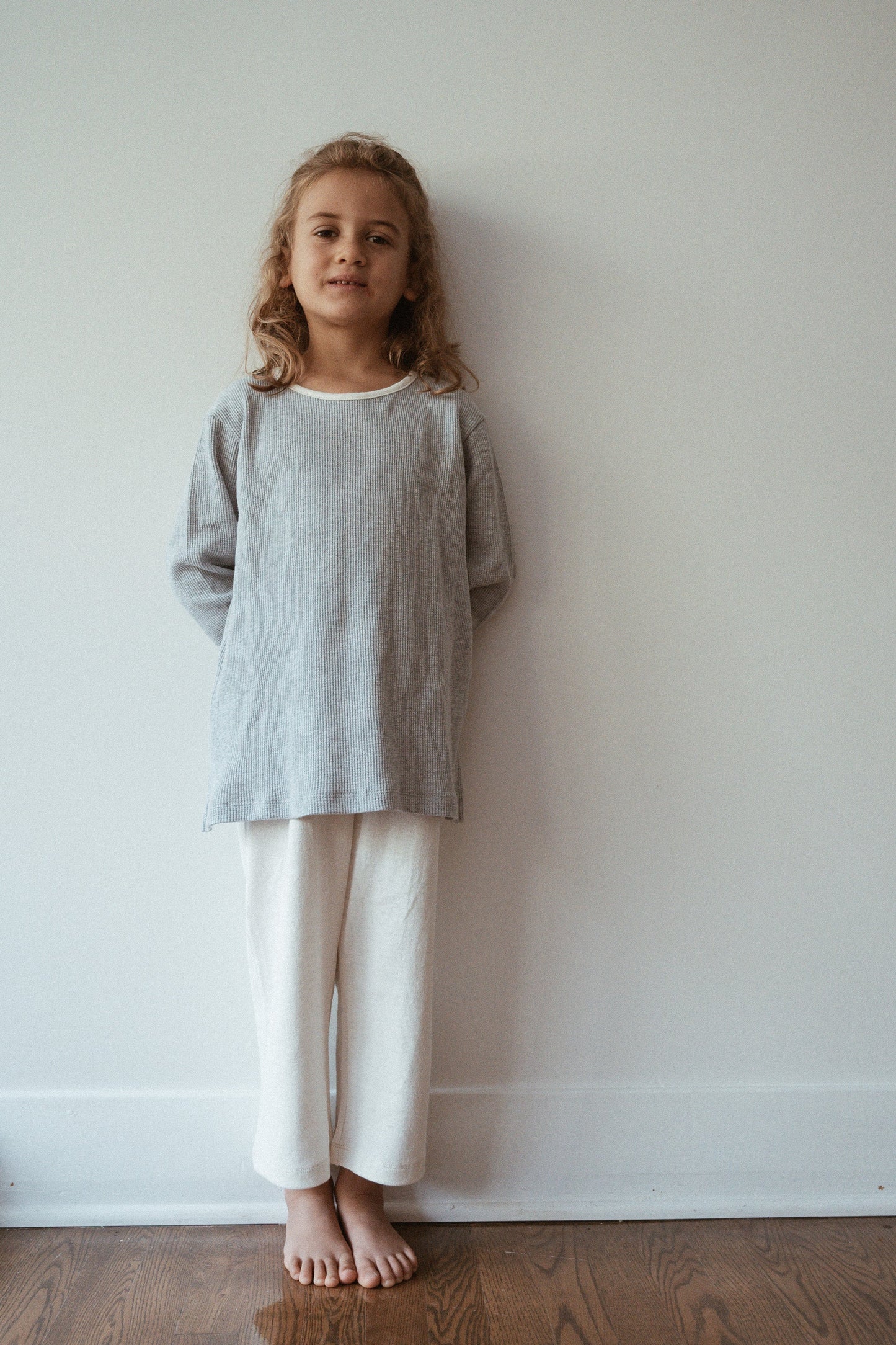 A young child stands indoors against a plain, light-colored wall in Calgary. They are wearing a light grey long-sleeve top and the ethically handmade white Resort Pant in Hemp from cabane childrenswear. The child, with light brown wavy hair, stands barefoot on a wooden floor, hands behind their back, gazing at the camera.