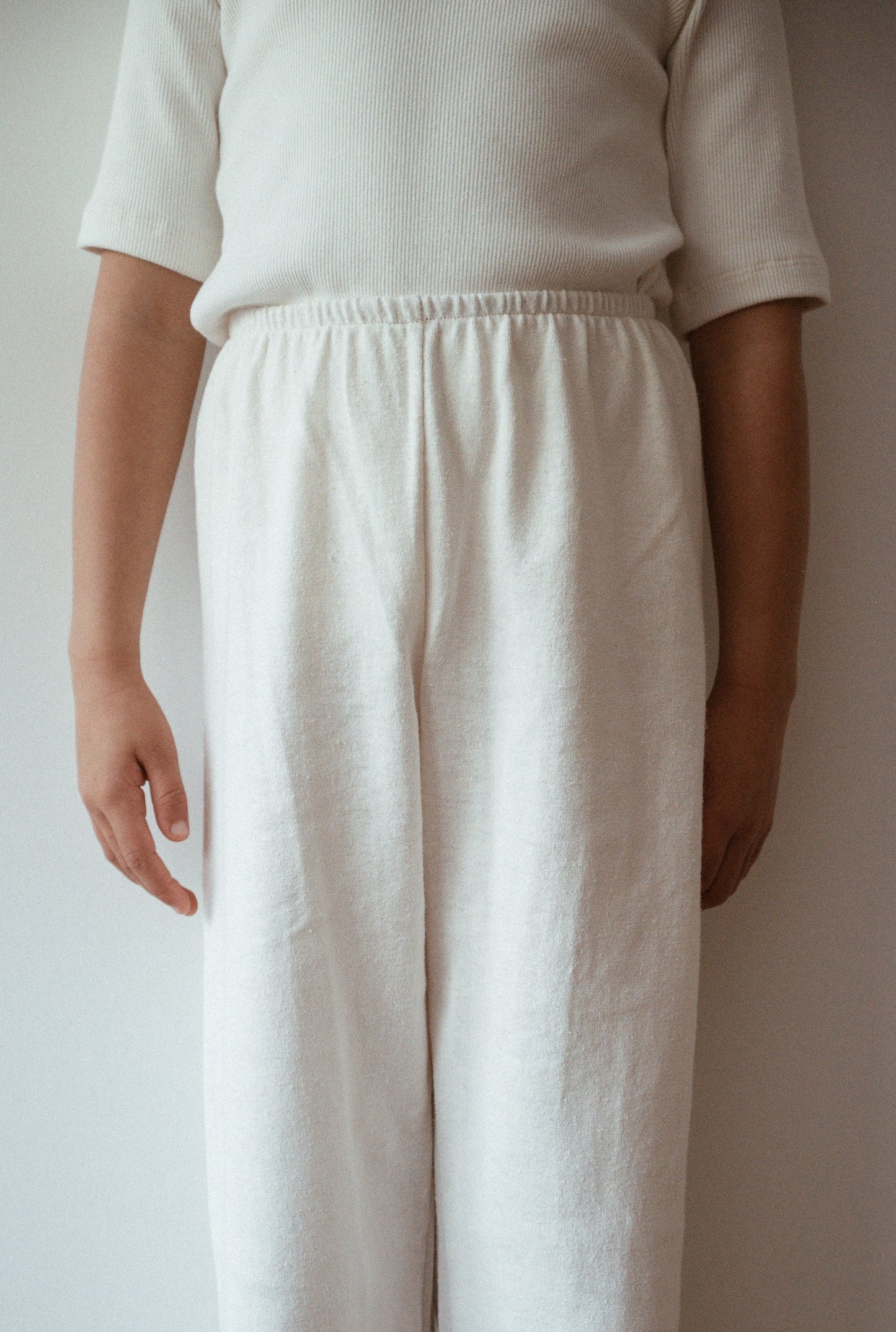 A child wearing off-white, textured RESORT PANT hemp pants and a matching ribbed shirt from cabane childrenswear stands in front of a light-colored wall. The child's arms hang loosely by their sides. The image captures only the child's torso and legs, highlighting the ethically handmade outfit.