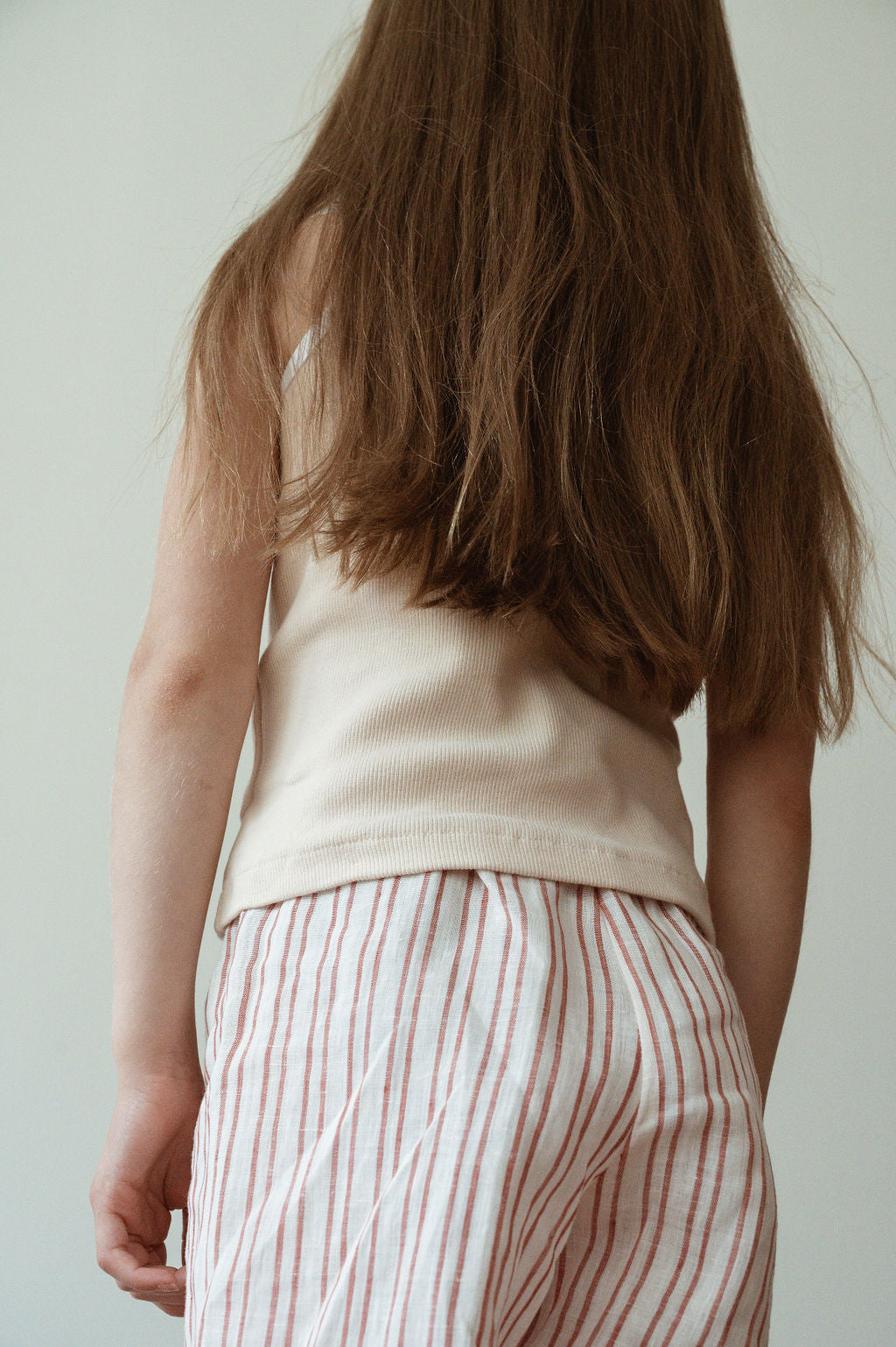 A young child with long, brown hair is pictured from the back, wearing a beige sleeveless top and ethically-made pink-and-white striped PER SE PANT loom linen pajama pants by cabane childrenswear. The background is plain and neutral.