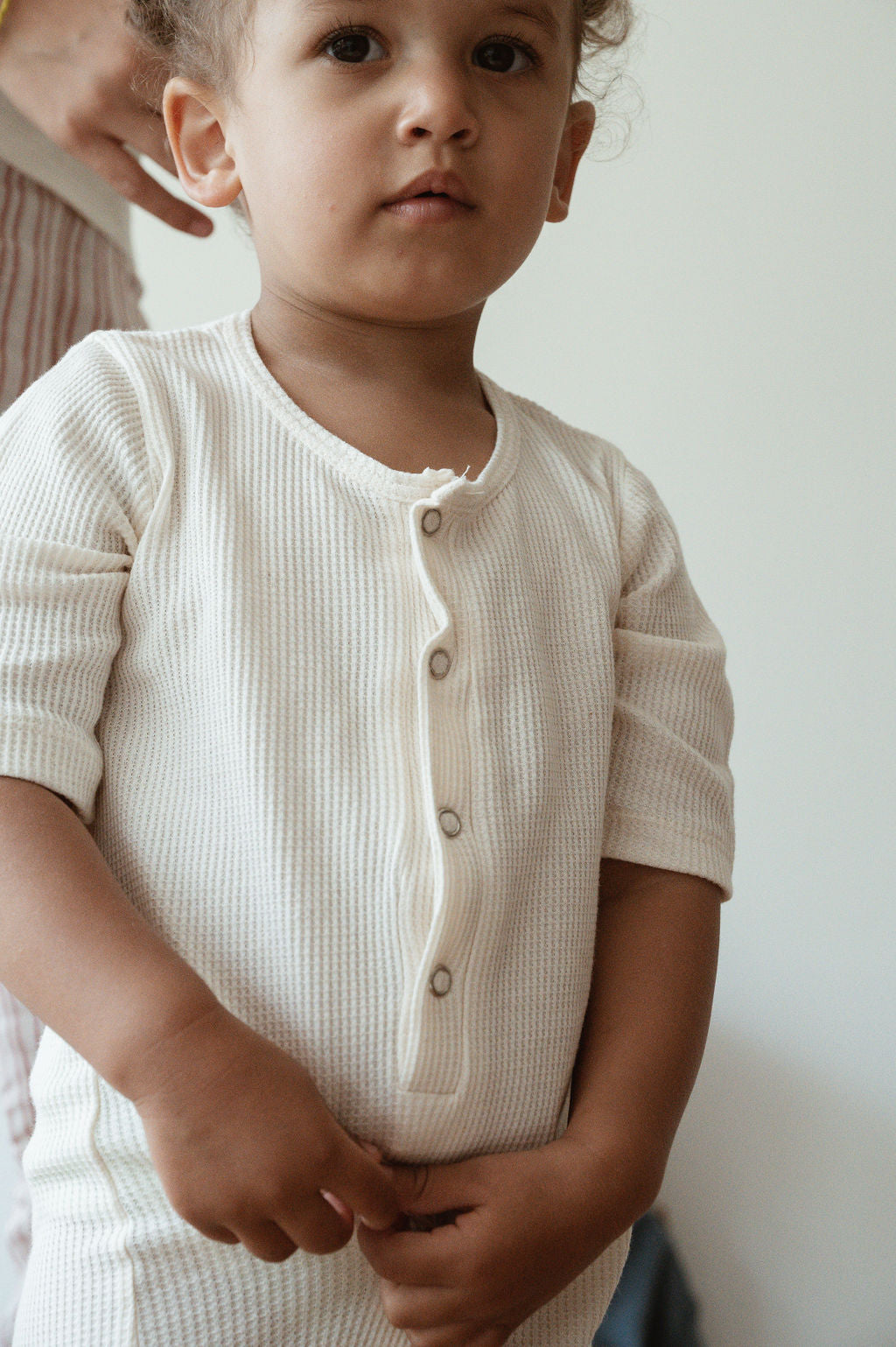 A young child with curly hair stands indoors, wearing the pre-order HIGH-NOON SUNSUIT from cabane childrenswear. The light-colored, buttoned, ribbed sunsuit is made from undyed organic cotton. The child holds their hands together and gazes slightly off-camera while another person is standing nearby, partially visible in the background.