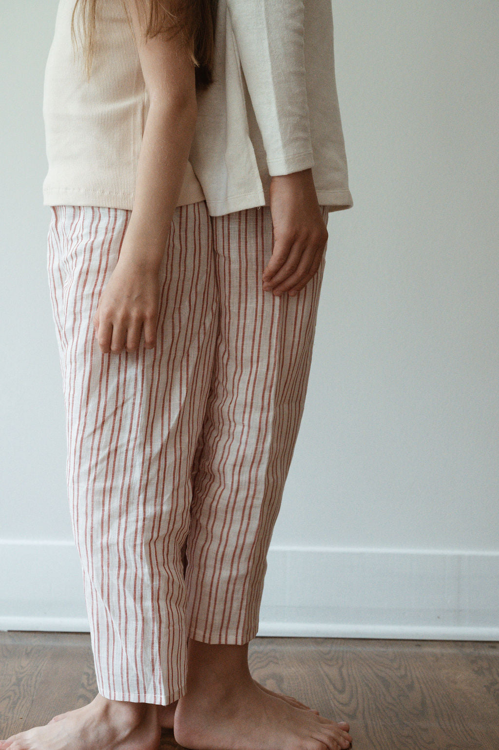 Two individuals dressed in light-colored, long-sleeved tops and relaxed PER SE PANT loom linen from cabane childrenswear are standing barefoot on a wooden floor. They are facing forward, but only their torsos and legs are visible in the image, highlighting the casually stylish look of their ethically made outfits.