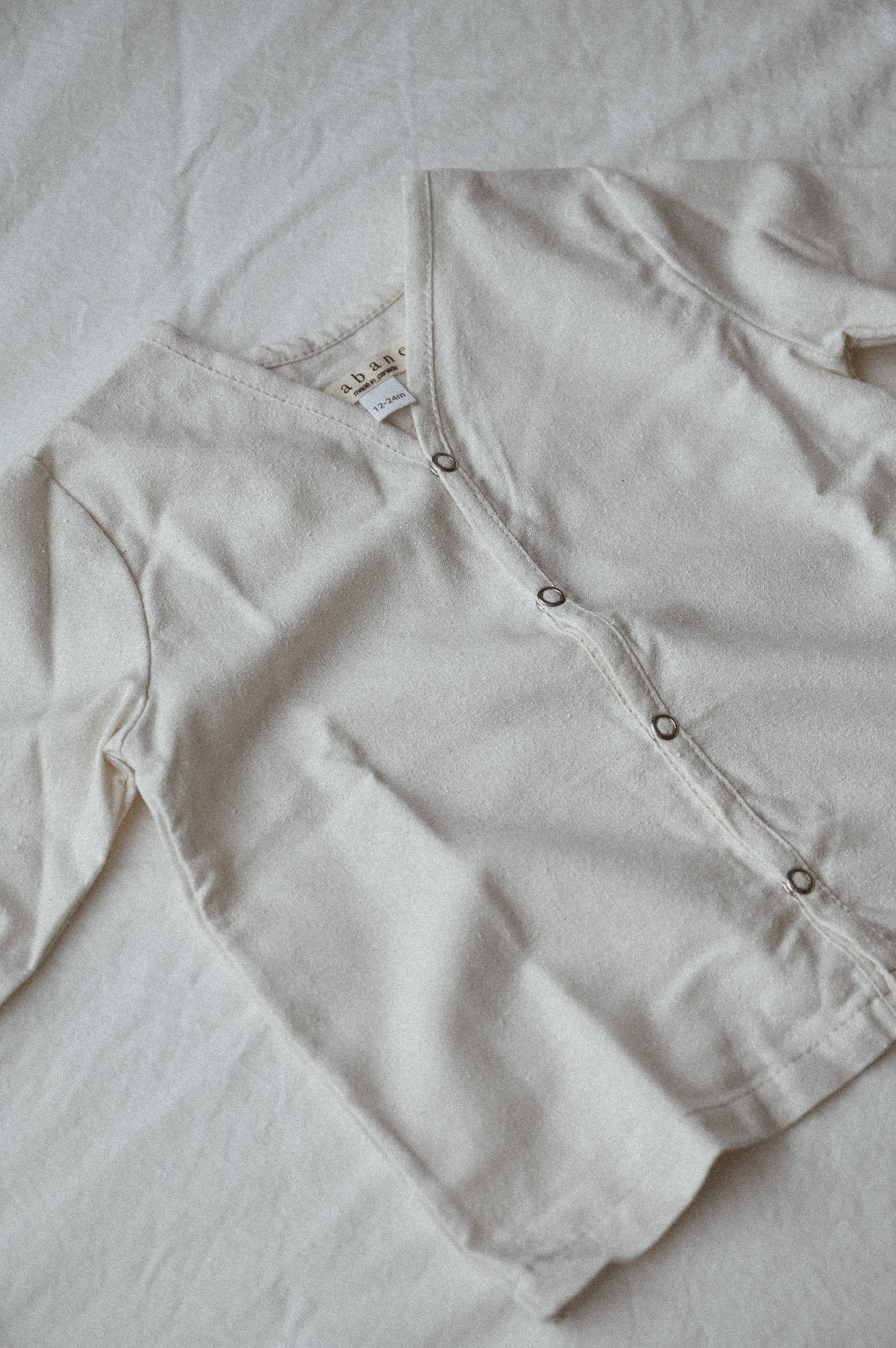 The TAKE CARE TOP raw silk by cabane childrenswear, a light-colored, long-sleeved, button-up shirt, is laid flat on a light-colored fabric surface. Ethically handmade in Calgary, this simple design features metal buttons and has a tag visible at the collar.