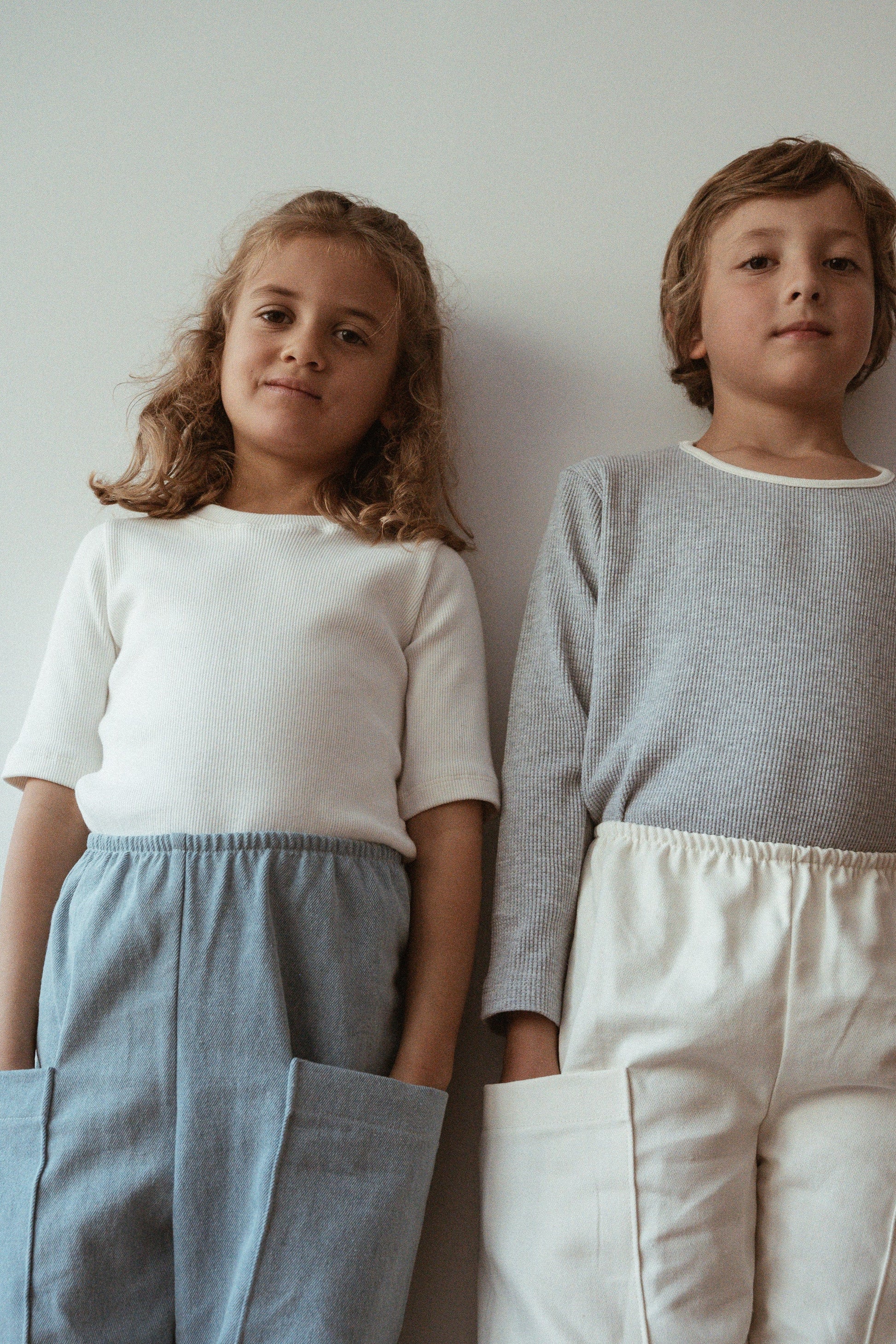 Against a light-colored wall, two children model outfits from cabane childrenswear's ethically handmade collection. The child on the left is dressed in a white top paired with the Pantalon D'Artiste in Denim, while the child on the right wears a gray top and white pants. With hands tucked into their pockets, both children gaze confidently at the camera.