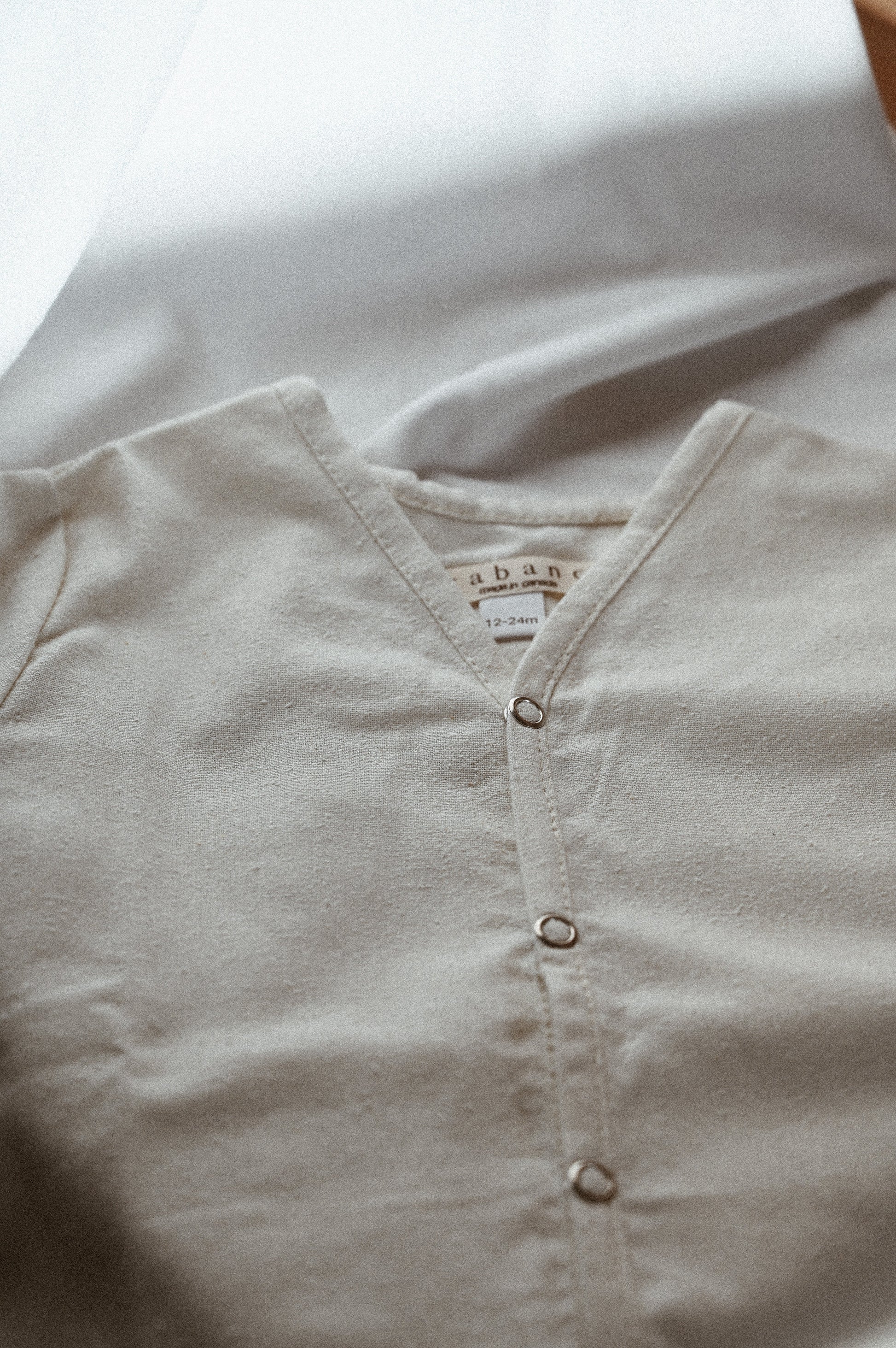 A close-up of a beige TAKE CARE TOP raw silk baby onesie with metal snap buttons, laid flat on a white surface. Ethically handmade in Calgary by cabane childrenswear, the tag inside the neck area indicates a size range of 0-24M. The fabric appears soft and comfortable for a baby.