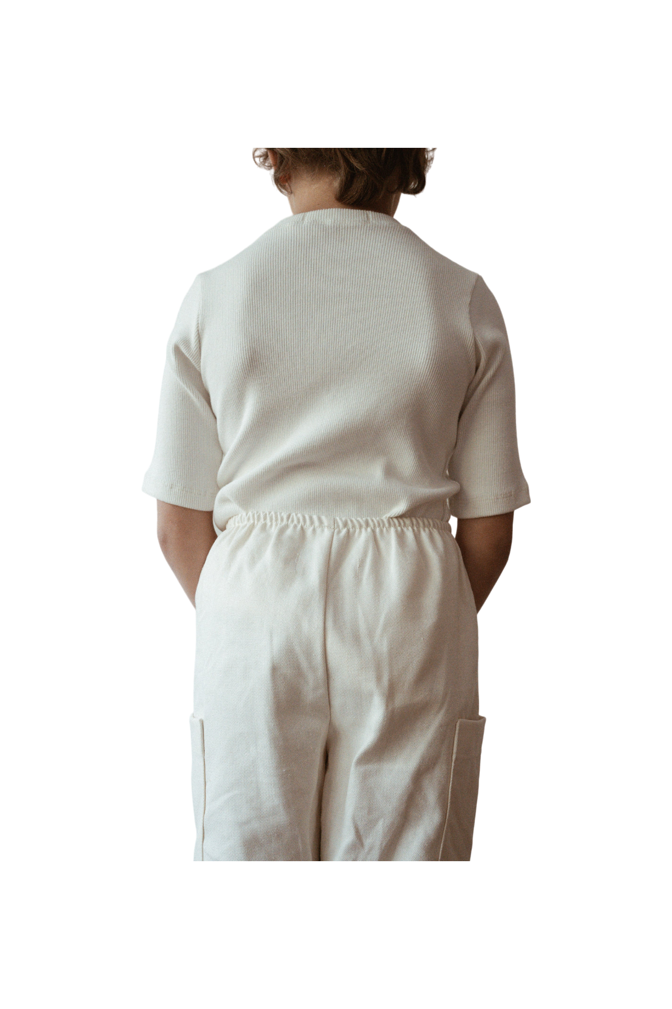 A child is standing with their back to the camera, wearing an ethically handmade white short-sleeved CLASSIC TOP baby + kid by cabane childrenswear and matching white pants. The child's hands are tucked behind their back, and the background is black, making the child the focal point of the image.