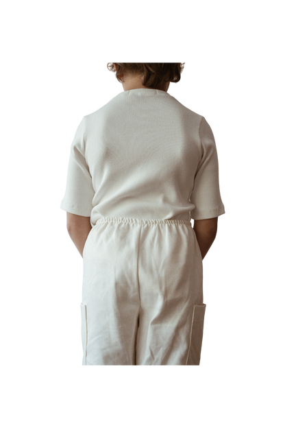 A child is standing with their back to the camera, wearing an ethically handmade white short-sleeved CLASSIC TOP baby + kid by cabane childrenswear and matching white pants. The child's hands are tucked behind their back, and the background is black, making the child the focal point of the image.