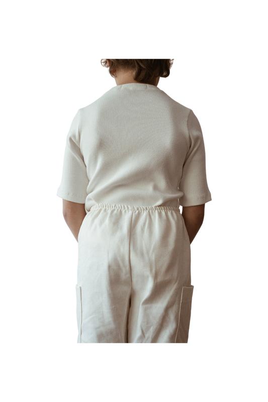 A child is standing with their back to the camera, wearing an ethically handmade white short-sleeved CLASSIC TOP baby + kid by cabane childrenswear and matching white pants. The child's hands are tucked behind their back, and the background is black, making the child the focal point of the image.