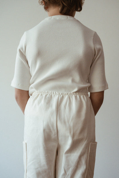 A person with shoulder-length hair stands with their back to the camera, dressed in a light-colored top from cabane childrenswear's Classic Top for Baby + Kid collection, paired with pants that have an elastic waistband. The backdrop is a simple off-white wall.