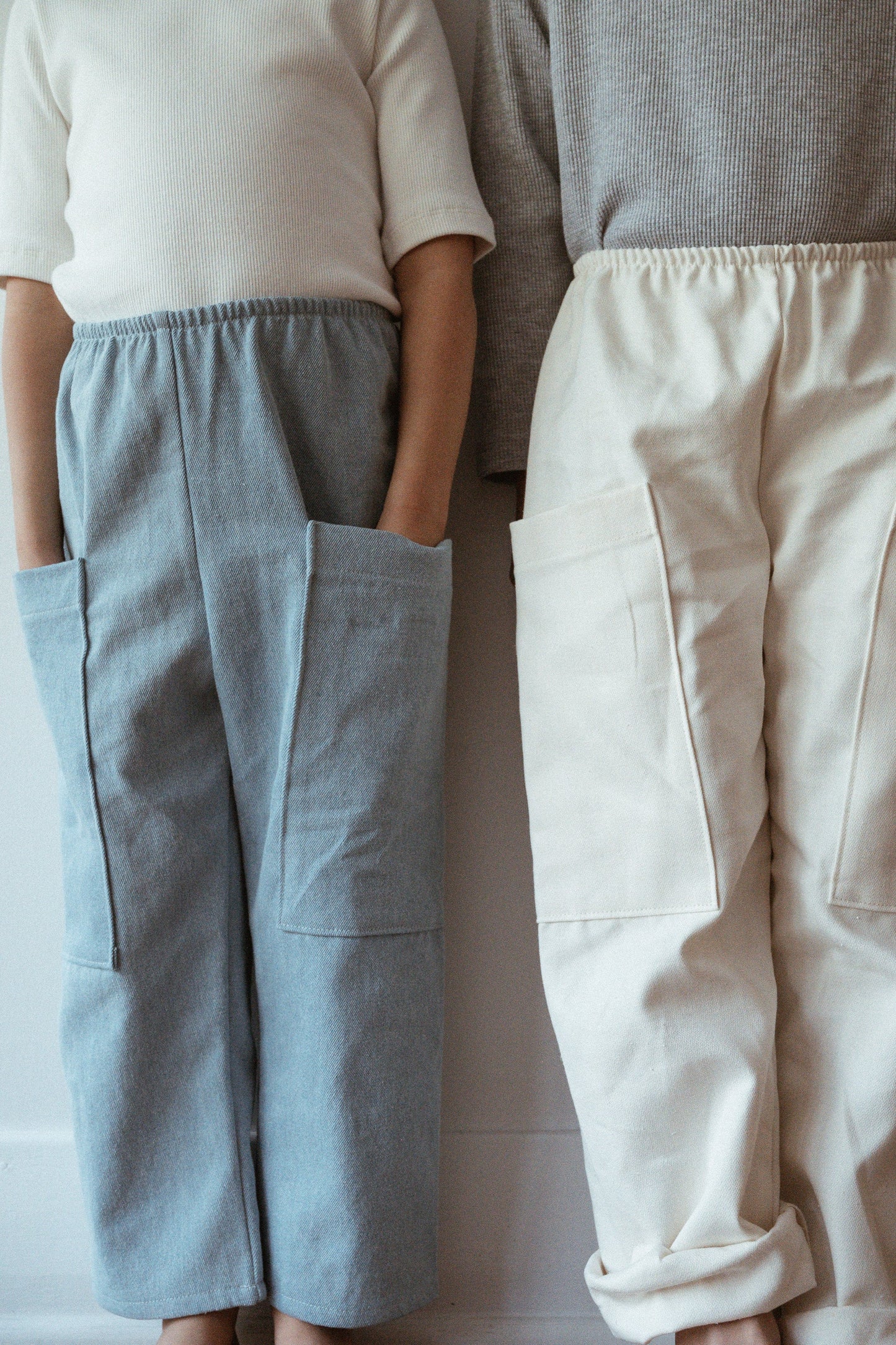 Two children stand side by side against a white wall, outfitted in ethically handmade long-sleeve tops and wide-leg pants featuring large pockets. One wears cabane childrenswear's Pantalon D'Artiste in Denim, while the other sports beige pants. Both have their hands tucked into their pockets, exuding a relaxed and casual vibe that perfectly captures Calgary's playful spirit.