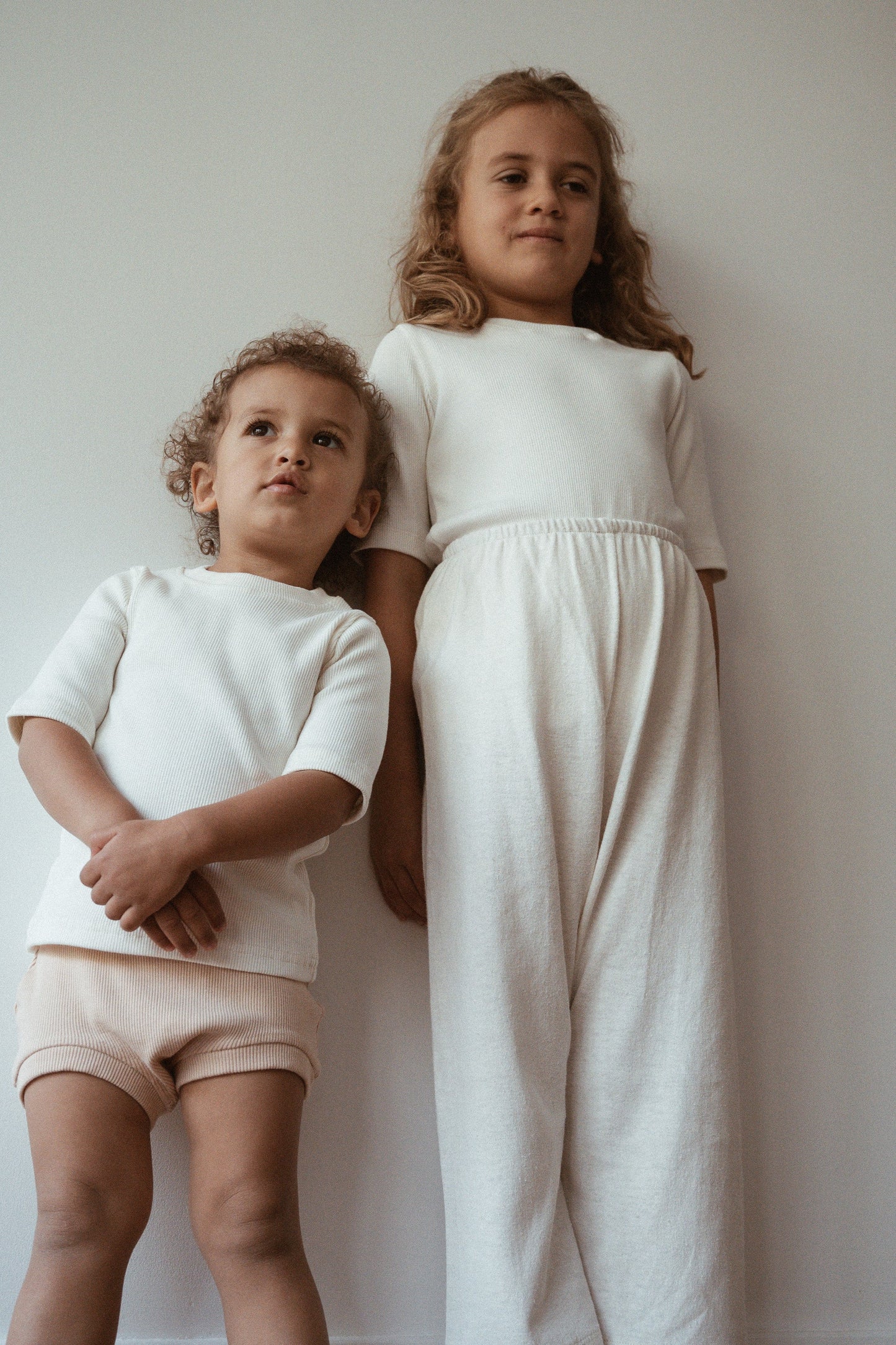 Two children stand against a plain white wall. The older child wears a cream dress, while the younger is dressed in an ethically handmade Classic Top for Baby + Kid by cabane childrenswear along with matching shorts. Both have curly hair and appear thoughtful in the soft, natural lighting.