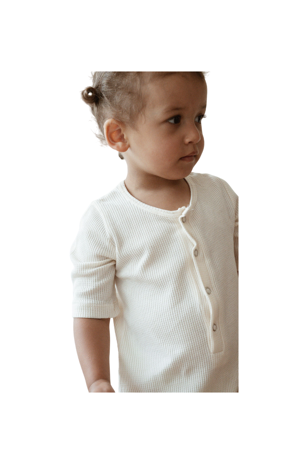 A young child with light brown hair is wearing the ethically handmade, ribbed, off-white HIGH-NOON SUNSUIT from cabane childrenswear. The undyed organic cotton fabric adds a touch of natural charm. The child is looking slightly to the right and has a neutral expression. The background is plain and dark.