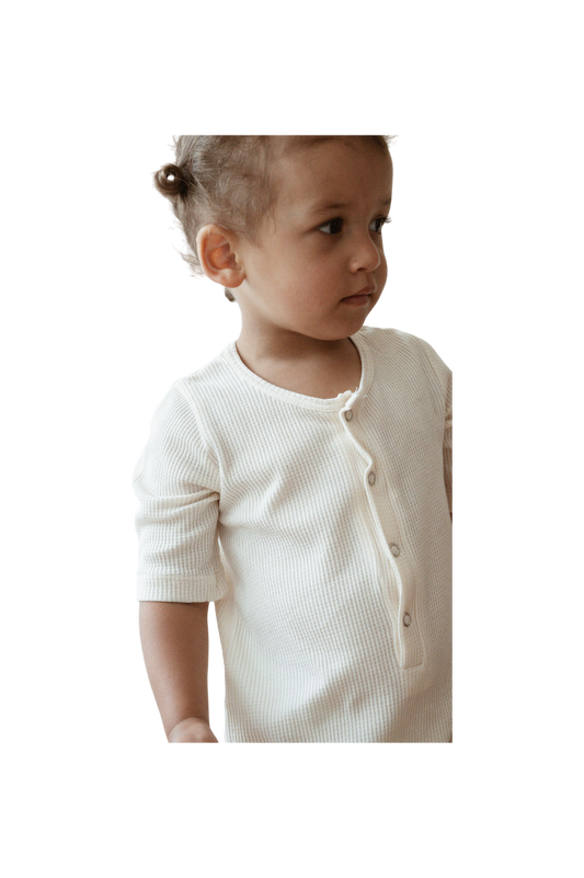 A young child with light brown hair is wearing the ethically handmade, ribbed, off-white HIGH-NOON SUNSUIT from cabane childrenswear. The undyed organic cotton fabric adds a touch of natural charm. The child is looking slightly to the right and has a neutral expression. The background is plain and dark.