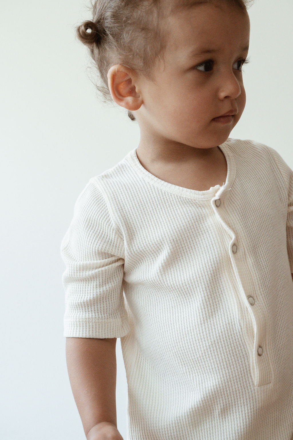 A young child with curly hair stands against a plain white background. The child is wearing the pre-order HIGH-NOON SUNSUIT by cabane childrenswear, made from light-colored undyed organic cotton in a long-sleeve waffle-knit design with buttons down the front. The ethically handmade shirt softly drapes as the child looks off to the side with a neutral expression.