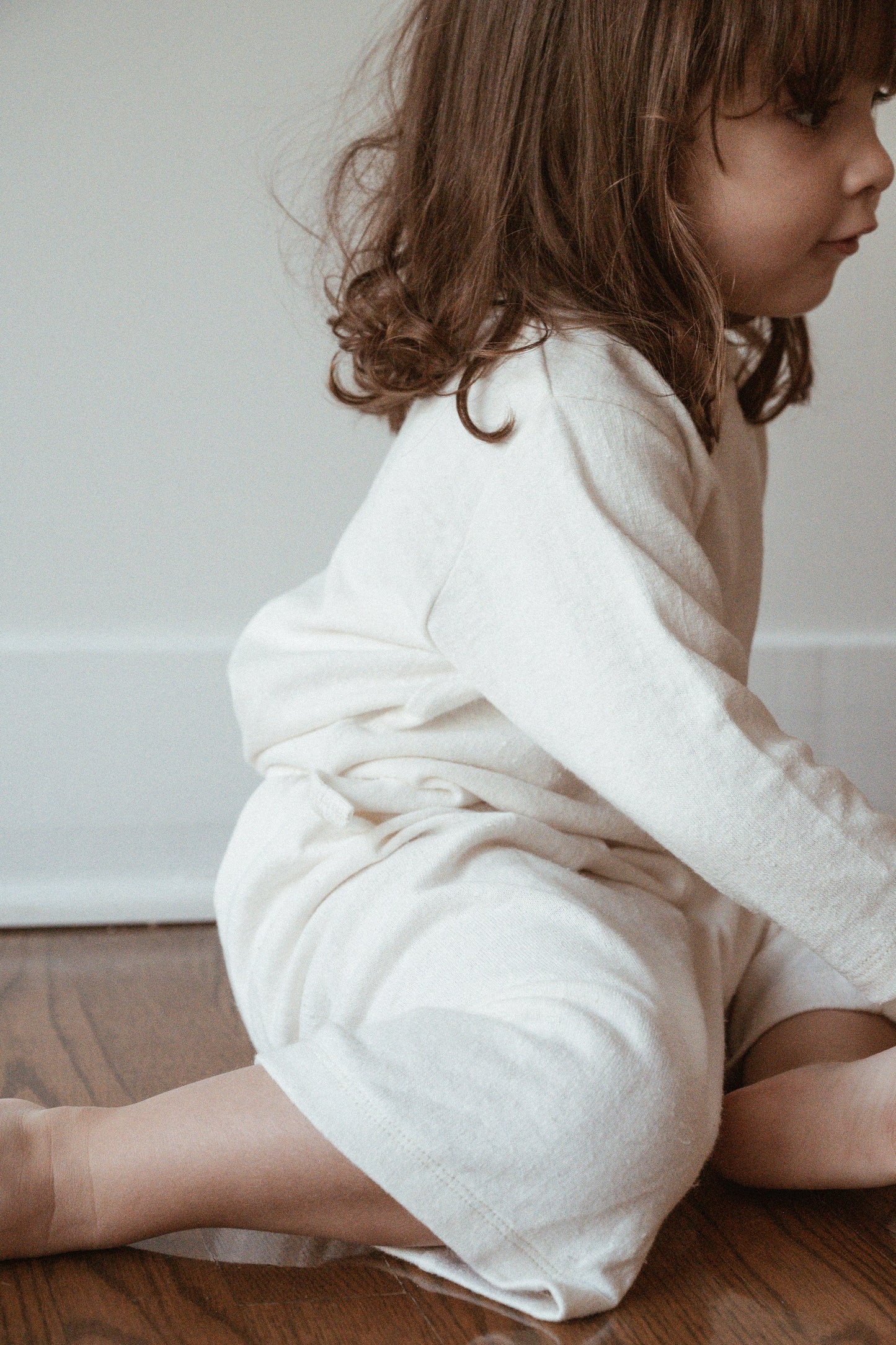 A young child with wavy brown hair, wearing the cabane childrenswear RESORT TOP hemp jersey in off-white organic cotton, sits on a wooden floor facing slightly to the right. The background is minimalistic, featuring a plain white wall.