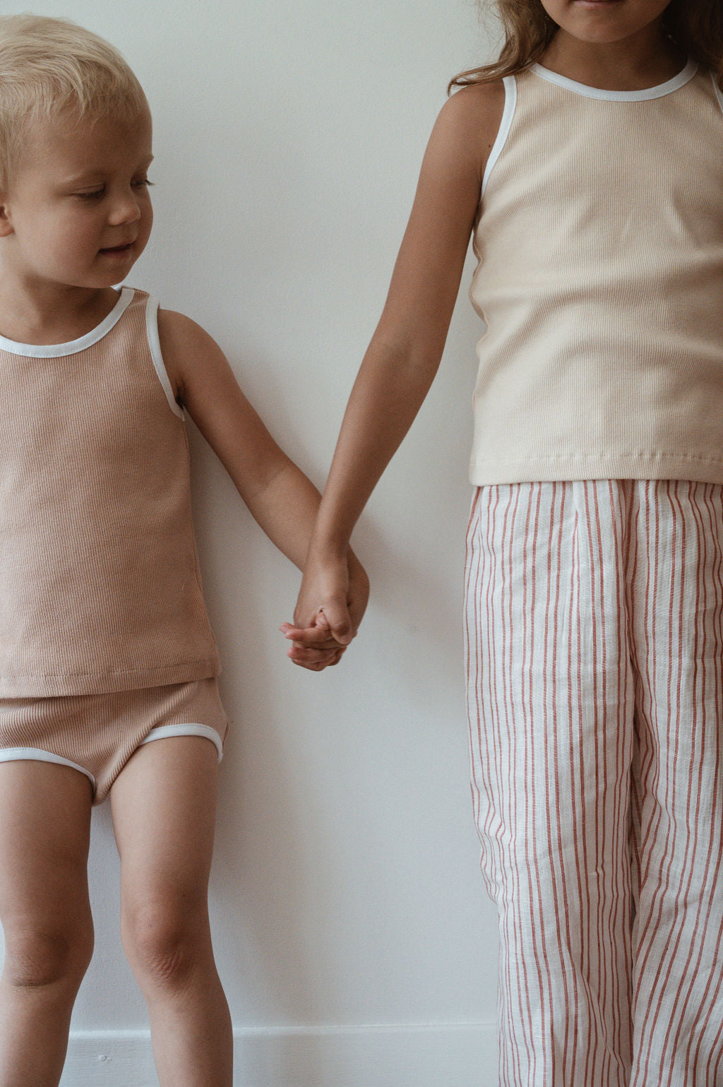 Two children holding hands stand against a white wall. The child on the left has light hair and wears a beige sleeveless top with matching shorts. The child on the right has longer hair and wears a beige sleeveless top paired with handmade, relaxed linen pants from cabane childrenswear's PER SE PANT loom linen collection.