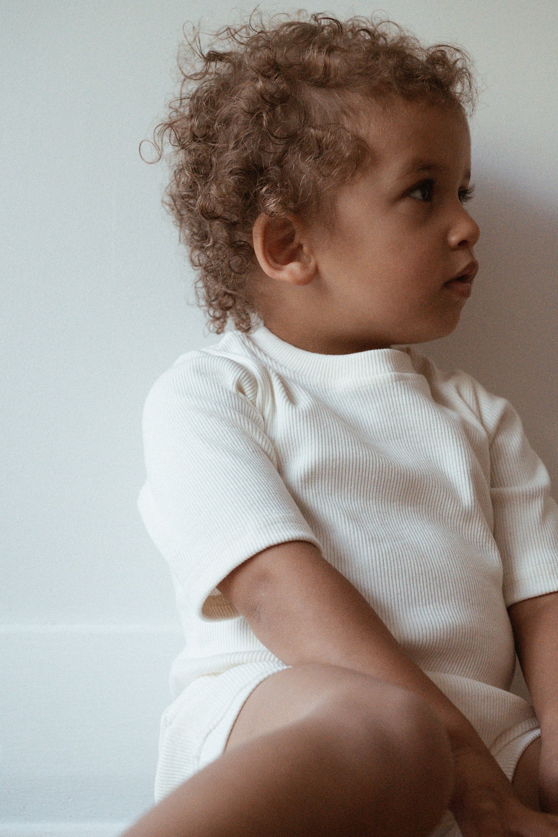 A young child with curly hair sits against a white wall, wearing a white ribbed short-sleeve onesie from the cabane childrenswear CLASSIC SET baby + kid. The child looks thoughtfully to the side, and the natural light highlights their profile.