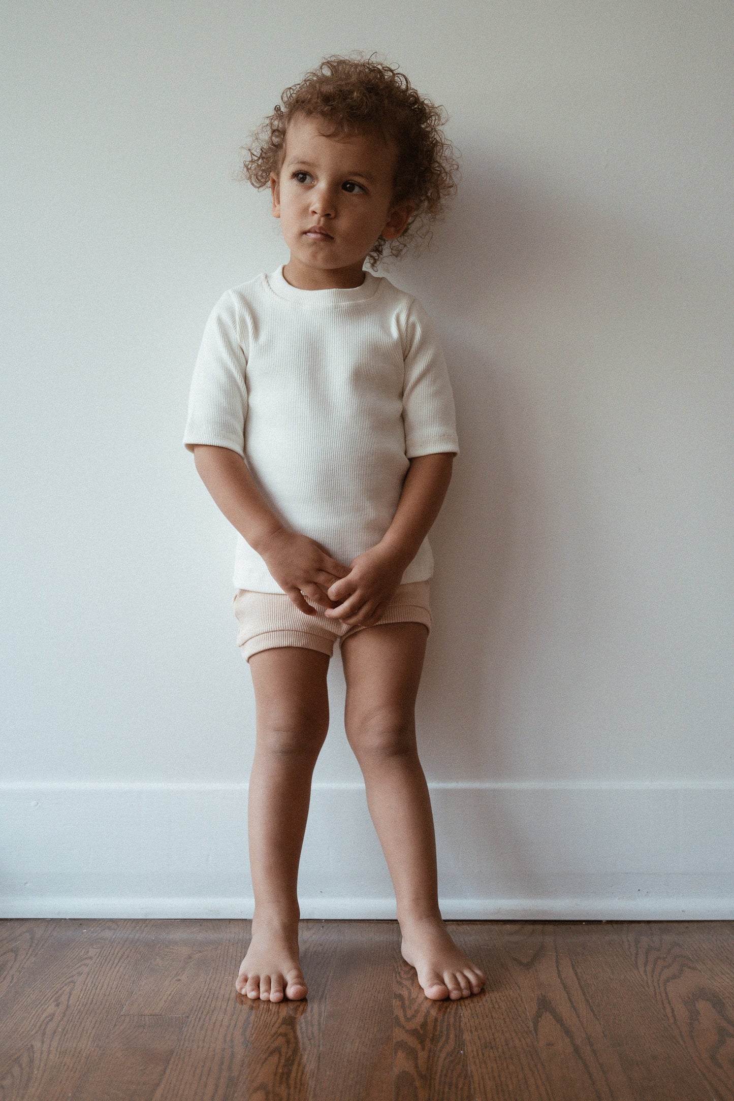 A young child with curly hair stands barefoot against a white wall, wearing the Classic Top for Baby + Kid from cabane childrenswear, paired with beige shorts. The child gazes thoughtfully to the side while standing on a wooden floor.