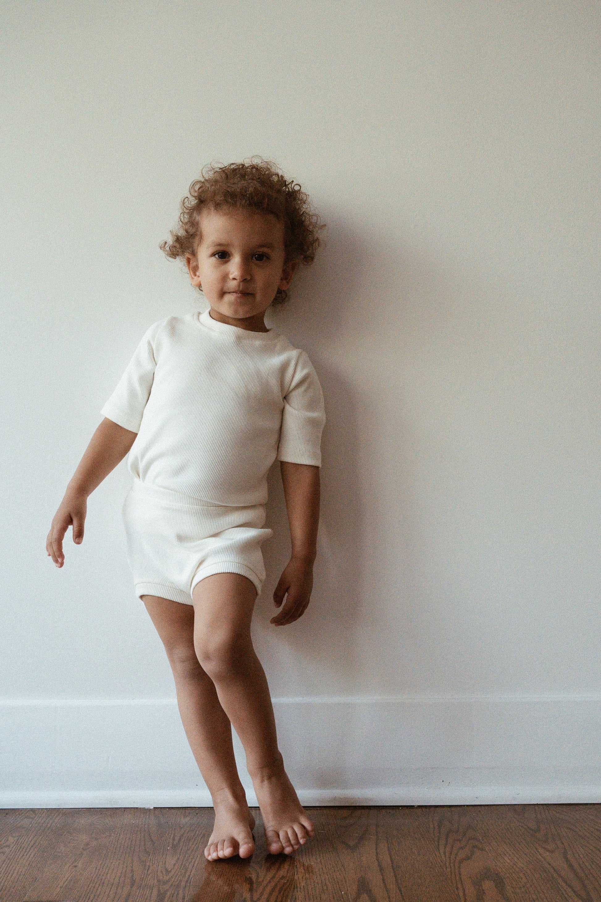 A young child with curly hair stands barefoot against a plain white wall, dressed in the CLASSIC SET baby + kid from cabane childrenswear. The white outfit, ethically handmade and featuring a rib texture, includes a short-sleeved top and matching shorts. The child looks into the camera with a relaxed posture, and the wooden floor is visible.