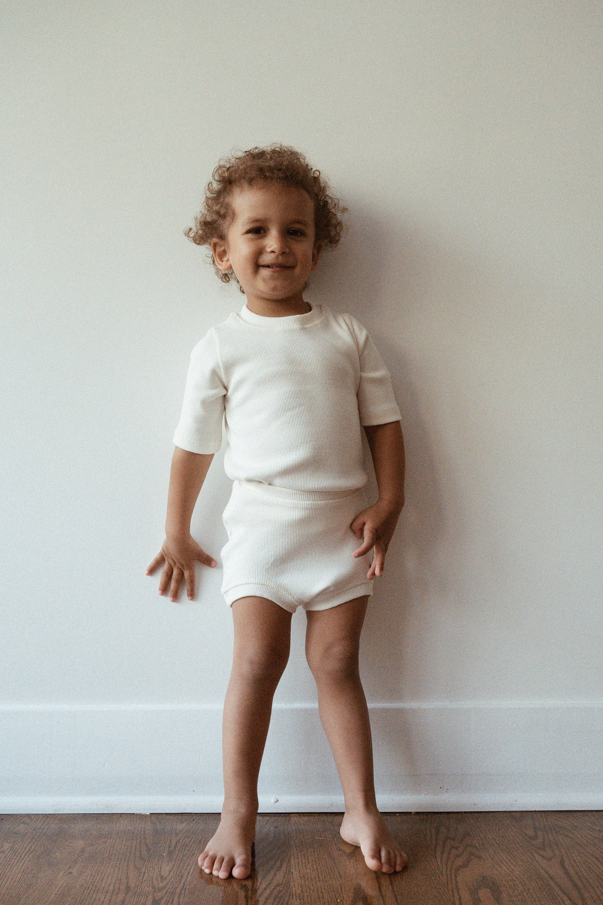 A young child with curly hair stands barefoot on a wooden floor against a plain white wall. The child is smiling and wearing an ethically handmade outfit with rib texture, consisting of a short-sleeve shirt and matching shorts from the CLASSIC SET baby + kid by cabane childrenswear. One hand rests casually on their hip while the other hangs by their side.