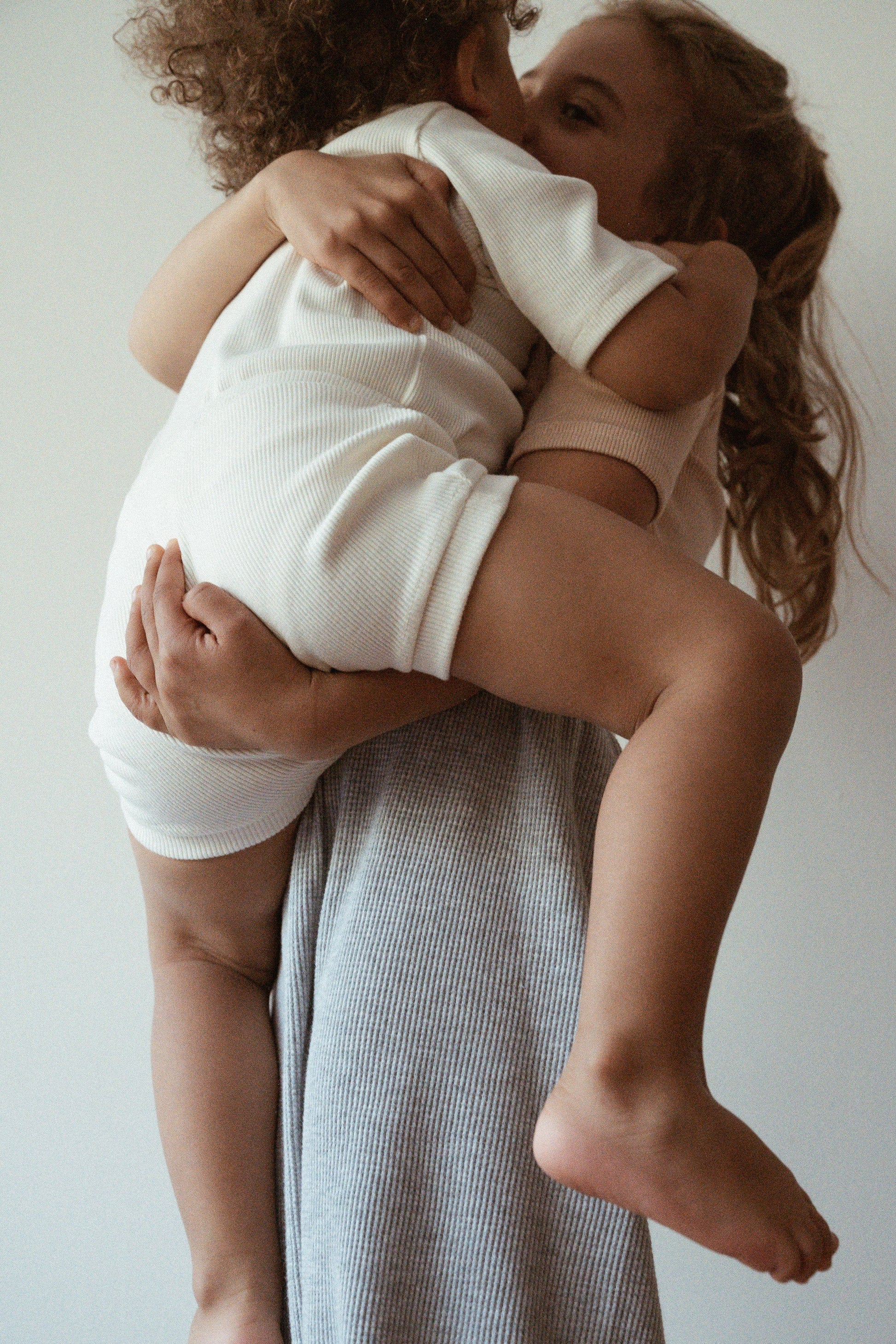 A young child dressed in a white outfit hugs a smaller child, who is also in a rib-textured white outfit, lifting them into the air. Both children are wearing warm smiles and showing affection against a plain background. Their embrace and body language convey strong sibling or familial love, reminiscent of the ethically handmade CLASSIC SET baby + kid by cabane childrenswear.