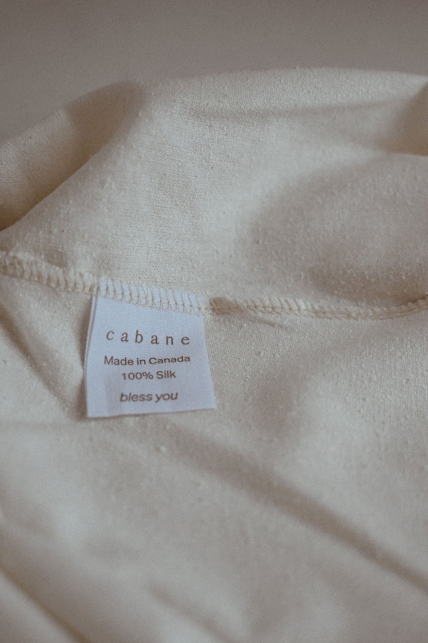 A close-up of a fabric label on a cream-colored silk garment. The label displays the brand name "cabane childrenswear," and notes that the item is the "TAKE CARE TOP raw silk" which is "ethically handmade in Calgary" from "100% raw silk fabric." Below the product details, the text "bless you" is printed.