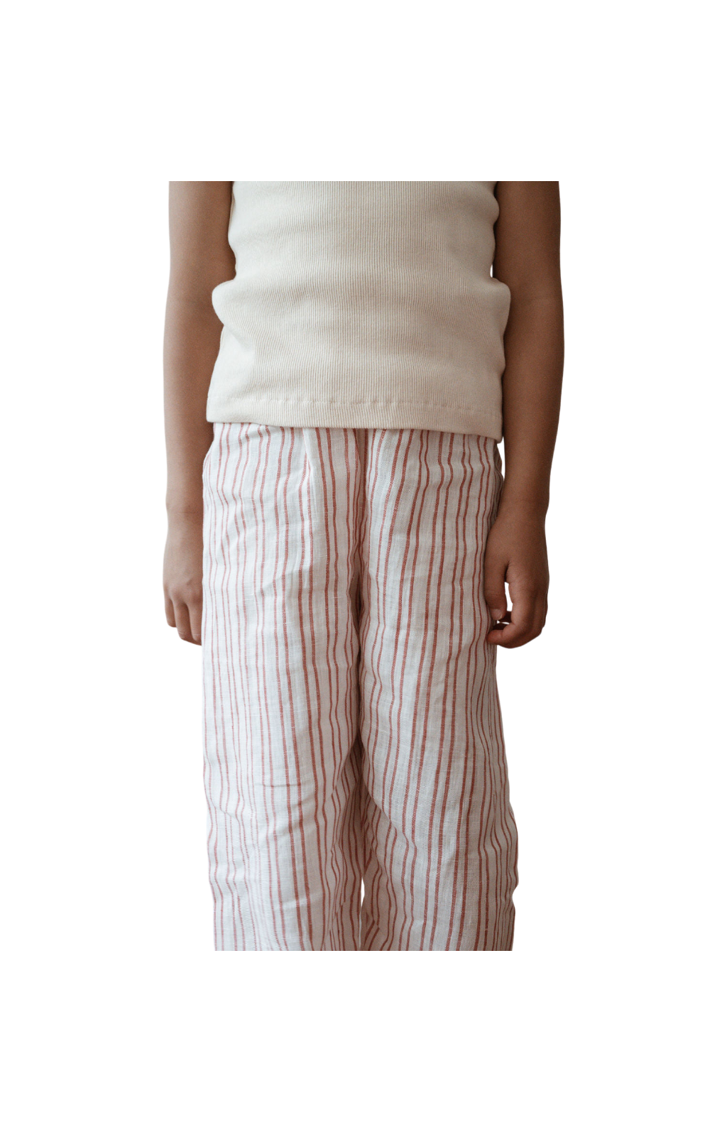 A person wearing a white sleeveless top and cabane childrenswear's PER SE PANT loom linen in a handmade pink and white striped pattern stands against a plain, dark background. The image only shows them from the neck down to their ankles.