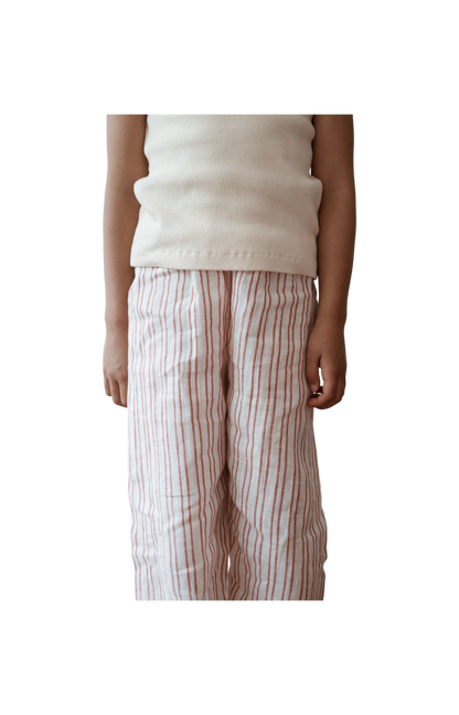 A person wearing a white sleeveless top and cabane childrenswear's PER SE PANT loom linen in a handmade pink and white striped pattern stands against a plain, dark background. The image only shows them from the neck down to their ankles.