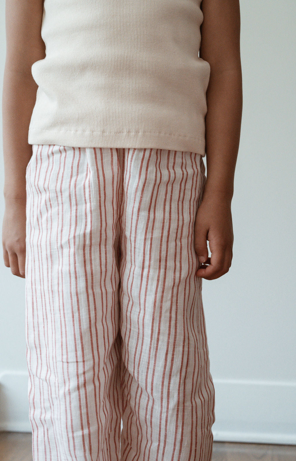 A child wearing a white tank top and PER SE PANT loom linen from cabane childrenswear, which features relaxed linen pants with white-and-red stripes, stands against a plain white background. The image is cropped to show the child from the neck to the knees, with arms relaxed at their sides. The outfit, featuring ethically made garments, exudes comfort and style.