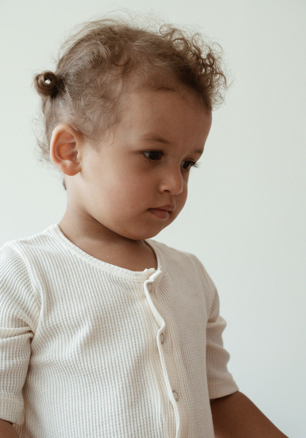 A young child with curly hair, tied in small ponytails, looks down thoughtfully while wearing the pre-order HIGH-NOON SUNSUIT from cabane childrenswear. The sunsuit is a light-colored ribbed shirt made from undyed organic cotton with buttons. The background is plain and minimalistic.
