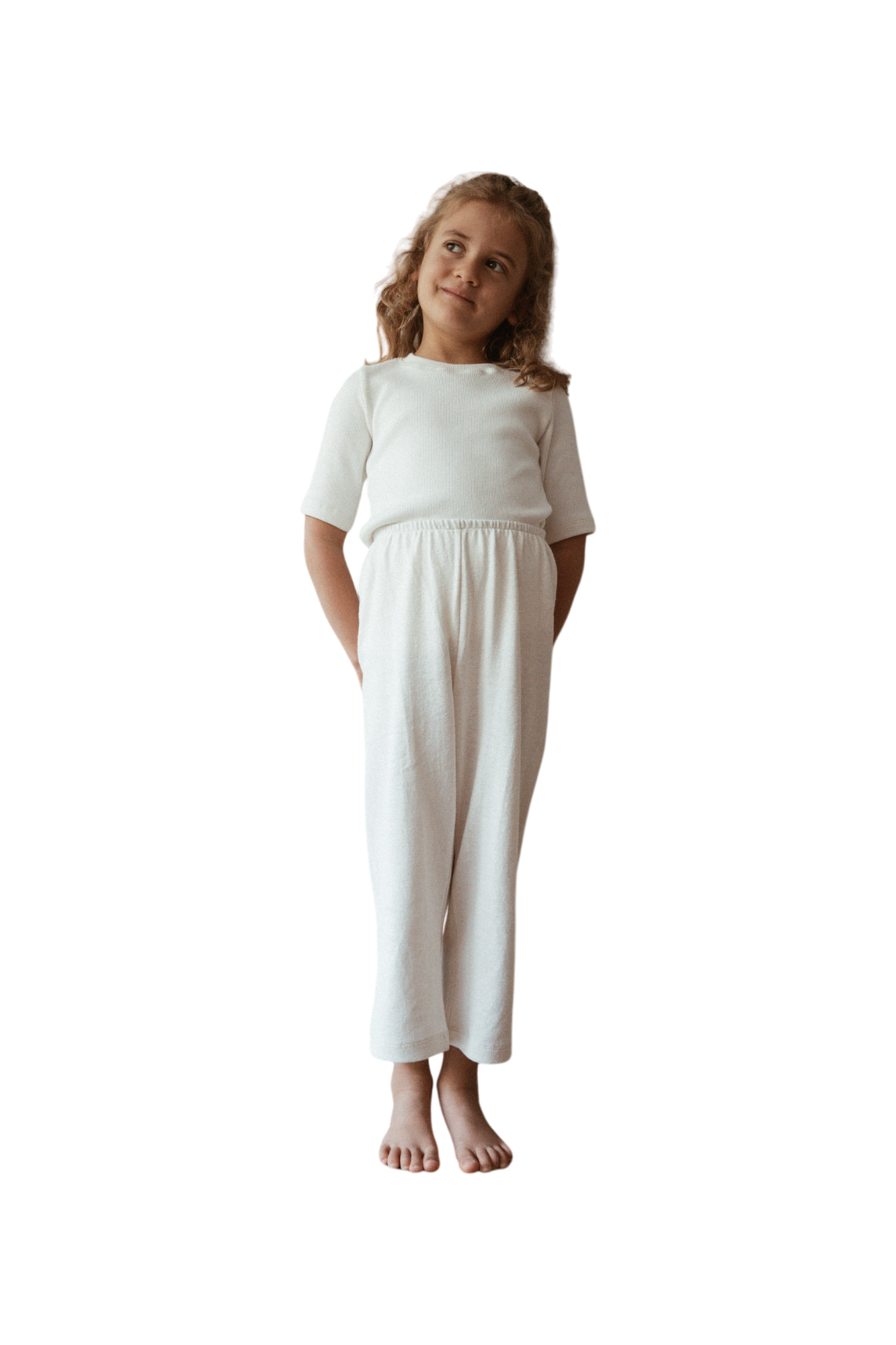 A young child with curly blonde hair stands barefoot, gazing thoughtfully to the side. The child is dressed in an ethically handmade white, short-sleeved top and matching RESORT PANT hemp from cabane childrenswear.